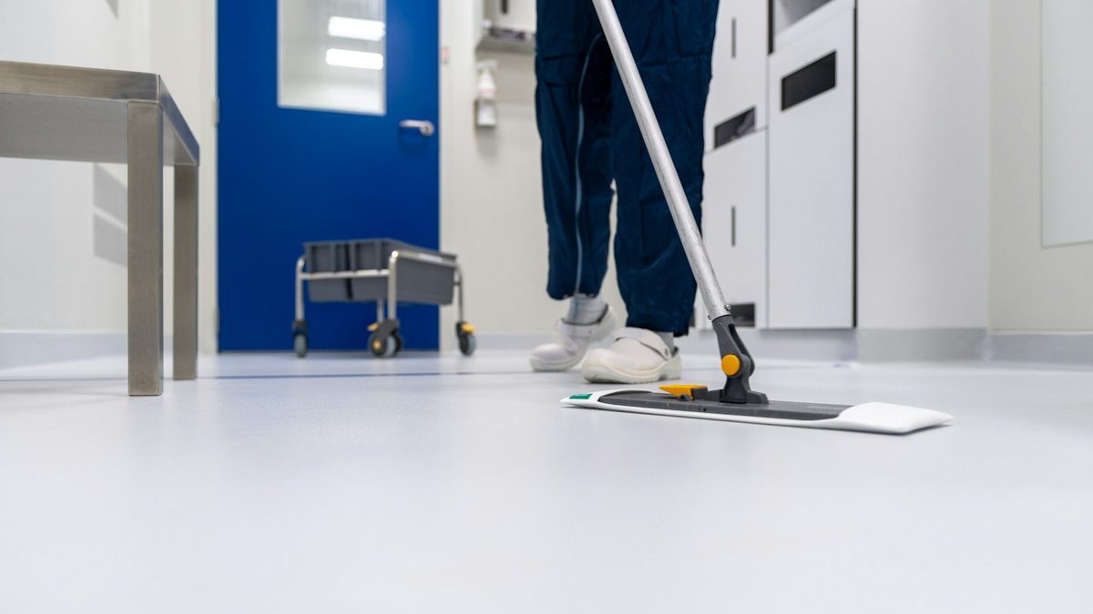 Reinigungskraft wischt den Boden eines hellen Klinikflurs mit einem Flachmopp